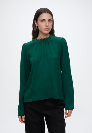 Femme aux longs cheveux foncés portant des boucles d’oreilles créoles dorées et un chemisier vert texturé à manches longues avec un pantalon noir sur fond uni.