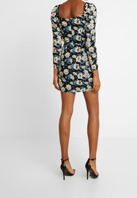 Kwiatowa sukienka na czarnym tle, z bufiastymi rękawami, kwadratowym dekoltem i dopasowanym krojem. Noszona z czarnymi sandałami na wysokim obcasie.