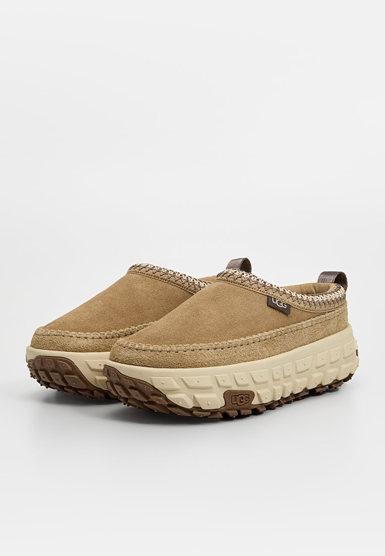 Zapatos de ante en tono beige, tipo slip-on, con suela de goma texturizada, costuras decorativas y una pequeña etiqueta con el logotipo en el lado.