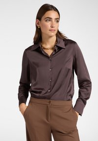 Camicia button-up in satin marrone con colletto e maniche lunghe, abbinata a pantaloni marroni. La camicia presenta una texture liscia e una vestibilità rilassata.