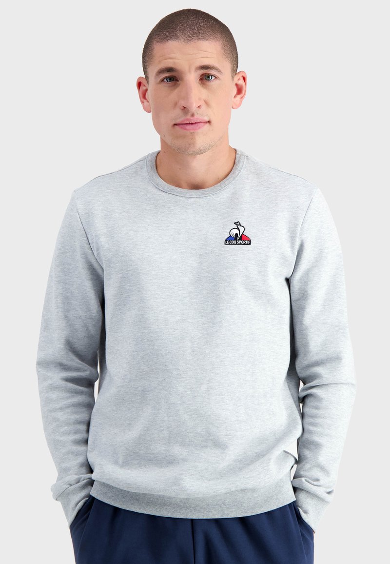 Sudadera gris claro con cuello redondo y mangas largas. Cuenta con un pequeño logo bordado en el pecho. Textura de tela suave.