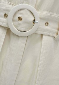 Cinturón de tela beige con hebilla redonda cubierta de tela y ojales metálicos sujeto a una prenda de algodón beige texturizada.