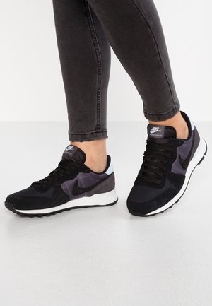 Personne portant des baskets Nike noires et gris foncé avec des lacets noirs et un jean slim gris foncé sur un sol et un fond blancs.