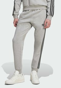 Grå bomulds sweatpants med sorte sidestrimler. Funktioner inkluderer elastisk talje og manchetter, parret med hvide sneakers. Adidas-logo på buksebenet.