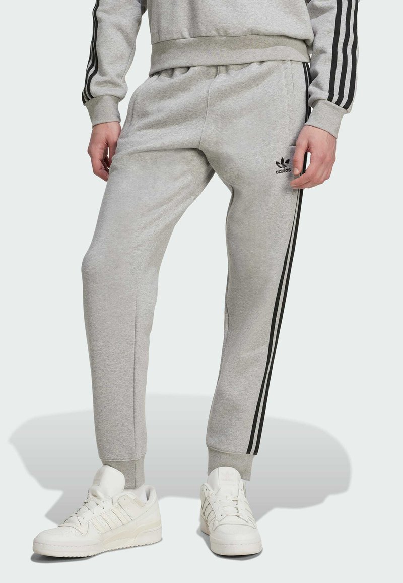 Grå bomulds sweatpants med sorte sidestrimler. Funktioner inkluderer elastisk talje og manchetter, parret med hvide sneakers. Adidas-logo på buksebenet.