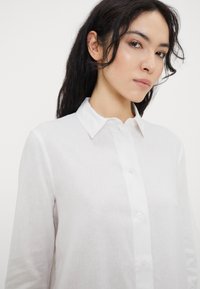 Camicia bianca a maniche lunghe realizzata in tessuto leggero, con colletto classico e un delicato motivo testurizzato.