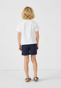 T-shirt bianco, pantaloni corti blu scuro e sandali grigi. Capelli ricci corti, visti da dietro, con design semplice e senza motivi visibili.