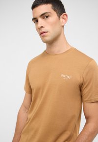 T-shirt marron à manches courtes en tissu lisse, avec un petit logo blanc "MUSTANG" sur le côté gauche de la poitrine et un col rond classique.