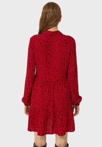 Robe rouge à manches longues, taille froncée, motif floral noir, et jupe à volants. Fabriquée en tissu léger, détail du col plat.