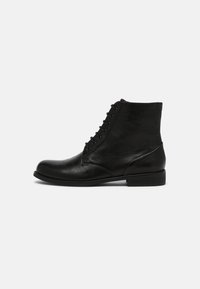Botín de cuero negro hasta el tobillo con diseño de cordones, punta redonda y un tacón bajo apilado. Presenta un acabado texturizado y un estilo minimalista.