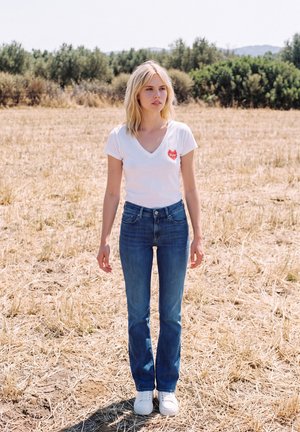 Une femme blonde portant un T-shirt blanc, un jean bleu et des baskets blanches se tient dans un champ sec, avec des arbres derrière elle.