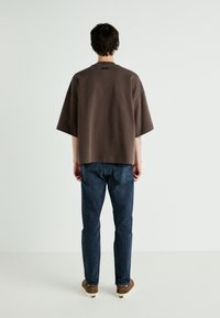 Barna, oversized pulóver kerek nyakkivágással, rövid ujjakkal és enyhén lejjebb vitt vállvonallal. Sötétkék farmernadrággal és barna cipővel viseli.