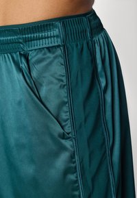Türkisblaue Sportshorts aus glänzendem Stoff, mit einem breiten elastischen Bund und seitlichen Taschen, aus einem glatten Material mit minimalen Nähten.