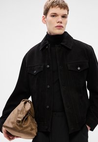 Melnā zamšādas jaka ar pogu aizdari un divām krūšu kabatām, valkāta virs melnas augstas apkakles džempera. Brūna ādas soma ar rāvējslēdzēju un rokturiem.