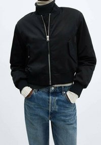 Černá zkrácená bomber bunda se zipem v přední části, měkkou texturou a dvěma bočními kapsami, nošená přes černý rolák a modré džíny.