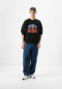 Een jonge man staat in een minimalistische ruimte, gekleed in een zwart sweatshirt met de tekst "ULTIMI ROMANTICI", losse blauwe jeans en witte sneakers.