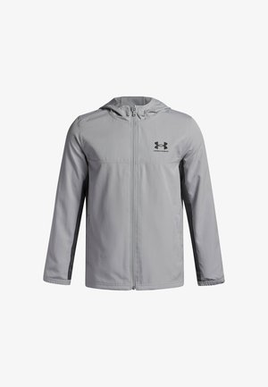 Chaqueta cortavientos de color gris claro, hecha de un suave tejido de nailon, con cierre de cremallera en la parte delantera, capucha y un logo negro en el pecho, con acentos oscuros en los laterales.