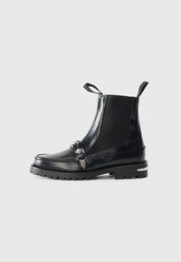Bottes noires en cuir à la cheville avec des panneaux élastiques sur les côtés, une sangle décorative avec des accents en métal, une semelle robuste et des tirettes pour un enfilage facile.