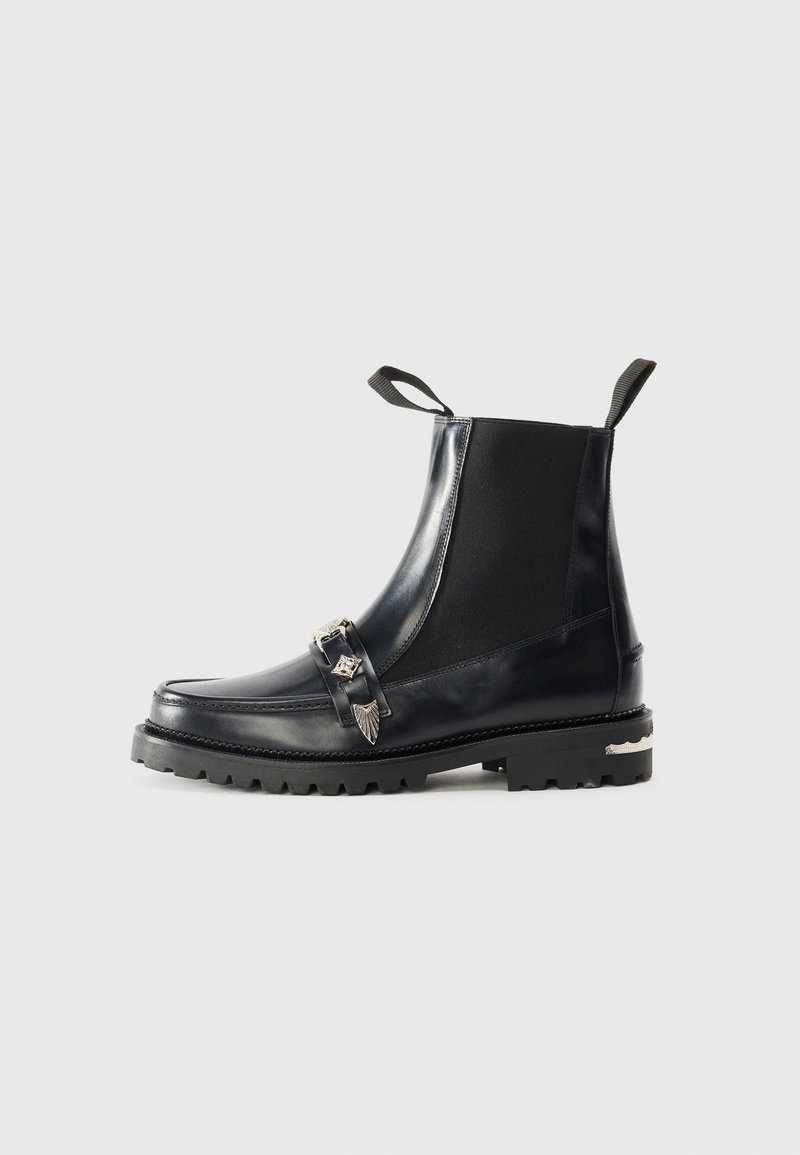 Bottes noires en cuir à la cheville avec des panneaux élastiques sur les côtés, une sangle décorative avec des accents en métal, une semelle robuste et des tirettes pour un enfilage facile.