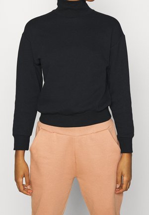Kvinde iført sort langærmet rullekravesweater og lysebrune bukser med hænder afslappet ned langs siderne mod en ensfarvet baggrund.