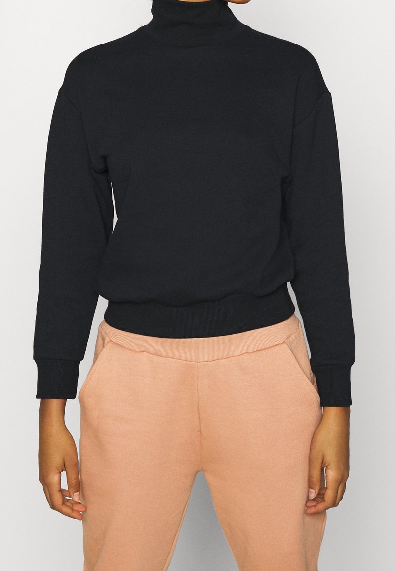Femme portant un pull à col roulé noir à manches longues et un pantalon marron clair, les mains détendues le long du corps, sur un fond uni.