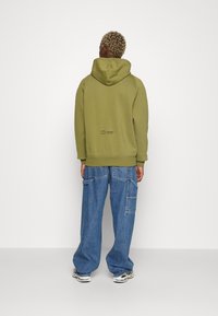 Sudadera con capucha de color verde oliva, con una capucha grande y puños acanalados, combinada con pantalones cargo de mezclilla azul claro de corte suelto que cuentan con múltiples bolsillos.
