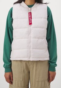 Helle rosa Steppweste mit einem Reißverschluss vorne, rundem Kragen, Seitentaschen und einem roten Etikett-Detail. Über einem grünen Langarmshirt getragen.