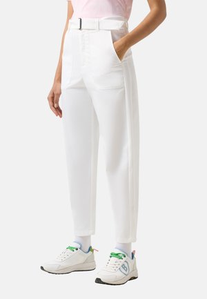 Person trägt hochgeschnittene weiße Hose mit großen Fronttaschen und Gürtel, kombiniert mit weißen Sneakers mit grünen Schnürsenkeln.