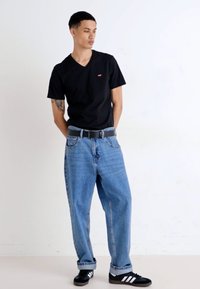 Tricou negru cu guler în V, blugi largi de culoare albastru deschis, centură gri și adidași negri cu dungi albe. Bărbatul stă cu mâinile pe șolduri.