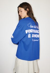 Blauwe oversized sweatshirt met witte tekst op de achterkant. De tekst luidt "FORMAGGIO È AMORE" en heeft een hartgrafiek. Zachte stof, relaxed fit.