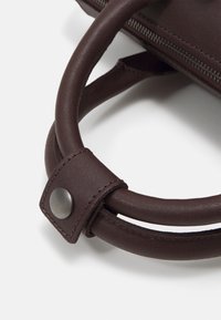 Gros plan de poignées de sac à main en cuir marron foncé avec bords cousus et détail de bouton-pression en métal sur fond blanc.