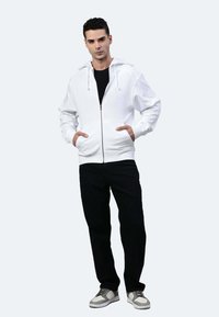 Weißer Zip-Hoodie mit Kordeln, Fronttaschen, getragen über einem schwarzen Shirt, kombiniert mit schwarzen Hosen und grauen Turnschuhen. Einfaches Design.