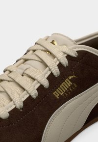Κοντινό πλάνο καφέ και μπεζ αθλητικού παπουτσιού Puma Tackle που δείχνει υφασμένα κορδόνια, υλικά από σουέντ και δέρμα, καθώς και το χρυσό λογότυπο Puma.