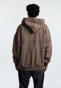 Felpa con cappuccio marrone, dal taglio rilassato, con grande testo stampato sul retro. Realizzata in un tessuto morbido con polsini e orlo a costine.
