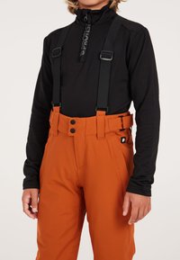 Pantaloni da sci arancioni isolati con bretelle nere regolabili, due bottoni frontali e una toppa con logo laterale. Abbinati a un top nero a maniche lunghe.