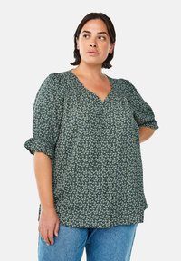 Blusa verde scuro con piccoli motivi floreali bianchi, scollatura a V e maniche corte a palloncino con bordo arricciato. Tessuto testurizzato.