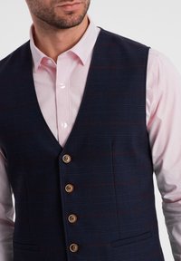 Gilet a quadri navy realizzato in tessuto texturizzato, con bottoni marroni e una camicia rosa chiaro in contrasto sotto.
