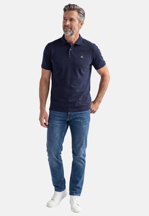 Homme aux cheveux gris et à la barbe portant un polo bleu marine, un jean bleu et des baskets blanches, debout devant un fond blanc.