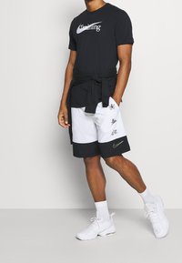 Hombre con camiseta negra de "Training", sudadera negra atada a la cintura, pantalones cortos blancos y negros con gráficos, calcetines blancos y zapatillas blancas.