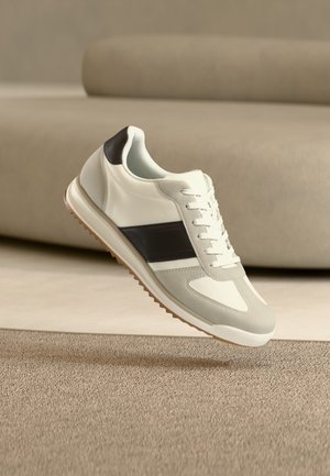 Baskets blanches avec une bande noire et une semelle marron flottant au-dessus d'un tapis beige dans une pièce aux tons neutres.