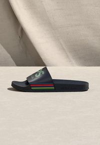 Σανδάλι slide σε ναυτικό μπλε με πολύχρωμες ρίγες στην πλευρά και έντονο πράσινο κείμενο στο λουρί, τοποθετημένο σε ανοιχτόχρωμη επιφάνεια.