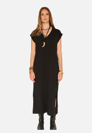 Femme debout portant une robe noire sans manches avec une fente latérale, des bottes noires, des colliers superposés et plusieurs bracelets aux deux poignets.