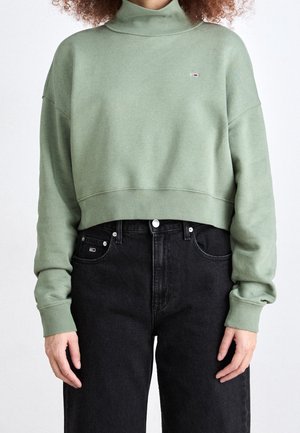 Person iført en salviegrøn cropped sweatshirt med rullekrave og højttaljede sorte jeans, stående mod en ensartet hvid baggrund.