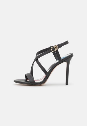 Black high-heeled sandal with thin crisscross straps, a small gold buckle, and a stiletto heel on a light background.