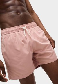 Bañador de color rosa claro hecho de material de secado rápido. Cuenta con una cintura elástica con un cordón blanco y costuras laterales sutiles.