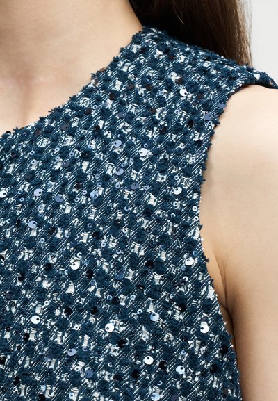 Αμάνικο σκούρο τιρκουάζ υφασμάτινο με υφαντά σχέδια και μικρές γυαλιστερές μαύρες παγιέτες, φορεμένο στο πάνω μέρος του σώματος κοντά στον λαιμό και τον ώμο.