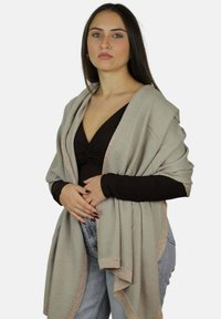 Młoda kobieta o długich ciemnych włosach, ubrana w czarną bluzkę, jasnoszary szal z błyszczącą lamówką oraz jasnoniebieskie jeansy, stojąca ze złączonymi rękami.