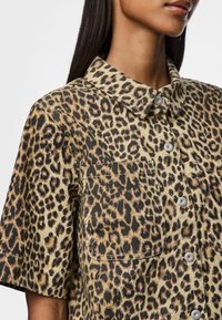 Camisa de botão com estampa de leopardo em tons de bege e preto, apresentando um colarinho, mangas curtas, bolso no peito e botões metálicos. Tecido texturizado.