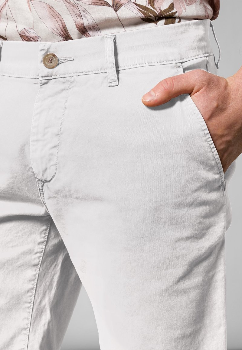 Main droite dans la poche d'un pantalon blanc avec une chemise fleurie rentrée, montrant un bouton et des passants de ceinture.