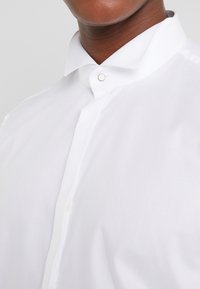 Primo piano di una persona che indossa una camicia bianca con colletto rigido e un singolo fermacravatta metallico visibile.